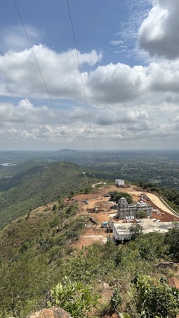 A scenic view of a hilltop with lush greenery. There is a construction site featuring a building with intricate architecture and machinery present. The landscape extends into the distance, with rolling hills and scattered clouds in a blue sky.