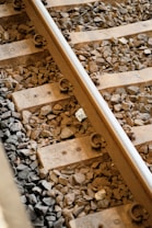 Railway tracks with wooden sleepers set on a bed of gravel. The metal rail is secured with fasteners, and the surrounding stones vary in size and color.