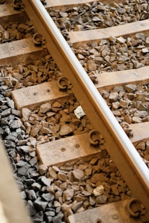 Railway tracks with wooden sleepers set on a bed of gravel. The metal rail is secured with fasteners, and the surrounding stones vary in size and color.