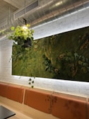 A modern interior space featuring a wall adorned with a lush vertical garden, filled with various types of green plants and small decorative elements. A suspended light fixture with intertwined leafy plants hangs from the ceiling, adding an organic touch to the industrial-style setting. The walls are made of white-painted bricks, while the seating area consists of simple orange padded benches.