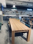 A modern cafeteria setting with a long wooden table surrounded by various chairs. The background features a tiled wall with decorative shelves and a neon sign that reads 'food4U'. Large overhead pendant lights add to a contemporary ambiance, and a small refrigerator is visible underneath the counter.