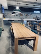 A modern cafeteria setting with a long wooden table surrounded by various chairs. The background features a tiled wall with decorative shelves and a neon sign that reads 'food4U'. Large overhead pendant lights add to a contemporary ambiance, and a small refrigerator is visible underneath the counter.
