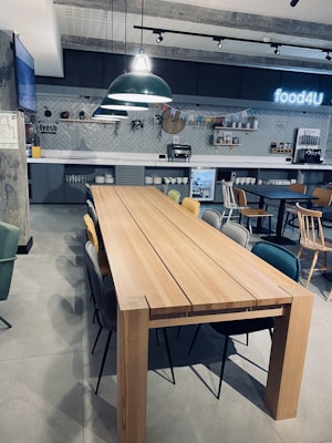 A modern cafeteria setting with a long wooden table surrounded by various chairs. The background features a tiled wall with decorative shelves and a neon sign that reads 'food4U'. Large overhead pendant lights add to a contemporary ambiance, and a small refrigerator is visible underneath the counter.