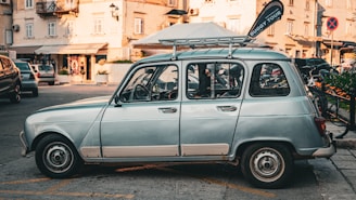 A vintage light blue car is parked on a street lined with buildings and shops. The car has a roof rack and a 'Buggy Tour' flag attached to it. There are bicycles and other parked cars in the background, and the scene takes place in what seems to be an urban area with a calm atmosphere.