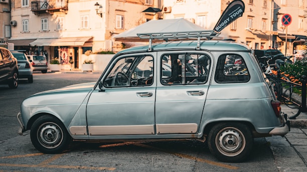 A vintage light blue car is parked on a street lined with buildings and shops. The car has a roof rack and a 'Buggy Tour' flag attached to it. There are bicycles and other parked cars in the background, and the scene takes place in what seems to be an urban area with a calm atmosphere.