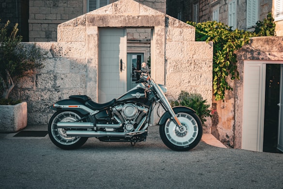 A classic motorcycle is parked in front of a rustic stone building, surrounded by potted plants. The building features an open door with large shutters and ivy cascading over parts of the stone wall.