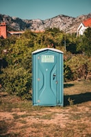 A blue portable toilet stands in the middle of a grassy area surrounded by lush green bushes. In the background, there are rocky mountains and houses with red-tiled roofs, under a clear blue sky.