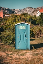 A blue portable toilet stands in the middle of a grassy area surrounded by lush green bushes. In the background, there are rocky mountains and houses with red-tiled roofs, under a clear blue sky.