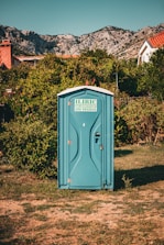 A blue portable toilet stands in the middle of a grassy area surrounded by lush green bushes. In the background, there are rocky mountains and houses with red-tiled roofs, under a clear blue sky.
