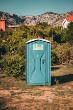 A blue portable toilet stands in the middle of a grassy area surrounded by lush green bushes. In the background, there are rocky mountains and houses with red-tiled roofs, under a clear blue sky.