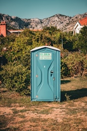 A blue portable toilet stands in the middle of a grassy area surrounded by lush green bushes. In the background, there are rocky mountains and houses with red-tiled roofs, under a clear blue sky.