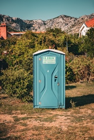 A blue portable toilet stands in the middle of a grassy area surrounded by lush green bushes. In the background, there are rocky mountains and houses with red-tiled roofs, under a clear blue sky.