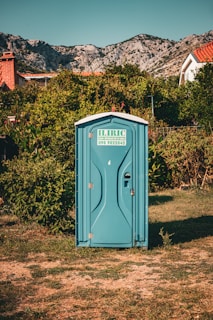 A blue portable toilet stands in the middle of a grassy area surrounded by lush green bushes. In the background, there are rocky mountains and houses with red-tiled roofs, under a clear blue sky.
