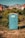 A blue portable toilet stands in the middle of a grassy area surrounded by lush green bushes. In the background, there are rocky mountains and houses with red-tiled roofs, under a clear blue sky.