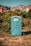 A blue portable toilet stands in the middle of a grassy area surrounded by lush green bushes. In the background, there are rocky mountains and houses with red-tiled roofs, under a clear blue sky.