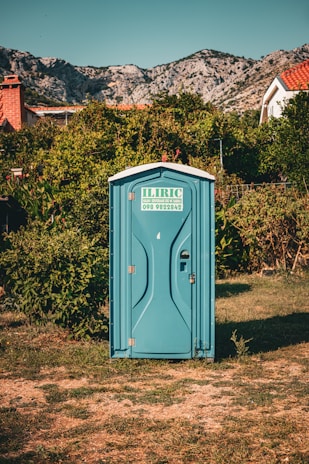 A blue portable toilet stands in the middle of a grassy area surrounded by lush green bushes. In the background, there are rocky mountains and houses with red-tiled roofs, under a clear blue sky.