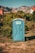 A blue portable toilet stands in the middle of a grassy area surrounded by lush green bushes. In the background, there are rocky mountains and houses with red-tiled roofs, under a clear blue sky.