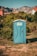 A blue portable toilet stands in the middle of a grassy area surrounded by lush green bushes. In the background, there are rocky mountains and houses with red-tiled roofs, under a clear blue sky.