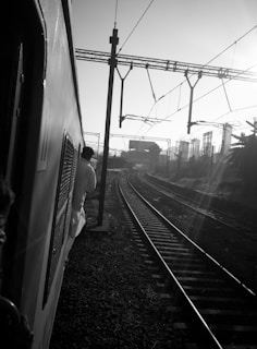 A black and white photograph captures a train journey with tracks extending into the distance. The image is taken from the side of the train with passengers visible leaning out and observing the scene. Overhead electric lines run parallel to the tracks, and structures, possibly buildings or industrial elements, are present in the background, with sunlight casting long shadows.