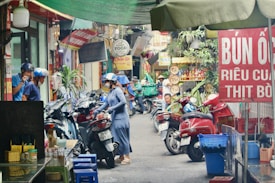 A bustling street scene featuring several motorbikes parked closely together. People are wearing helmets and appear engaged in various activities. Stalls and shops with vibrant signage are visible, offering food and beverages. The overall atmosphere is lively and colorful with various goods on display.