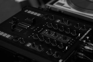 Close-up view of a DJ mixer featuring various knobs, sliders, and buttons for controlling music. The mixer has a sleek, dark design with clear labels and is connected to multiple cables, indicating its use in a professional audio setup.