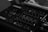 Close-up view of a DJ mixer featuring various knobs, sliders, and buttons for controlling music. The mixer has a sleek, dark design with clear labels and is connected to multiple cables, indicating its use in a professional audio setup.