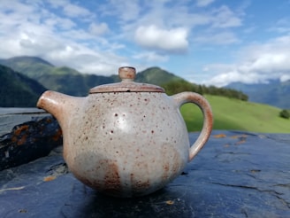 A ceramic teapot with a speckled texture is placed on a dark rock surface. In the background, a scenic view of rolling green hills and a blue sky with scattered clouds is visible.