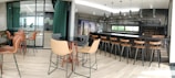 A modern bar and dining area featuring a sleek black counter with wooden bar stools. The space has contemporary decor with large windows allowing natural light to fill the room. There are round tables with comfortable chairs throughout, and the flooring consists of light wood panels. Industrial-style pendant lights hang from the ceiling.