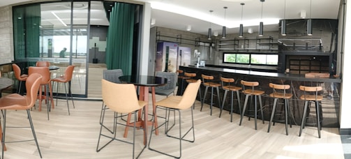 A modern bar and dining area featuring a sleek black counter with wooden bar stools. The space has contemporary decor with large windows allowing natural light to fill the room. There are round tables with comfortable chairs throughout, and the flooring consists of light wood panels. Industrial-style pendant lights hang from the ceiling.