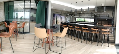 A modern bar and dining area featuring a sleek black counter with wooden bar stools. The space has contemporary decor with large windows allowing natural light to fill the room. There are round tables with comfortable chairs throughout, and the flooring consists of light wood panels. Industrial-style pendant lights hang from the ceiling.