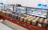 An array of assorted cakes is neatly arranged on a wooden table in front of a glass display case. The display case, filled with various desserts and pastries, is set against a white tiled wall. Blue and white balloons provide a festive touch in the background.
