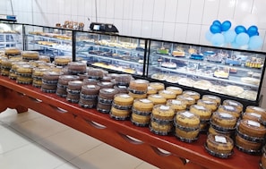 An array of assorted cakes is neatly arranged on a wooden table in front of a glass display case. The display case, filled with various desserts and pastries, is set against a white tiled wall. Blue and white balloons provide a festive touch in the background.
