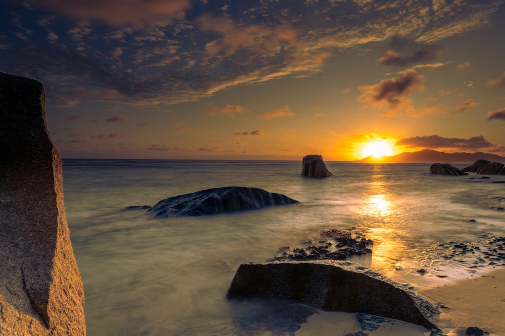 the sun is setting over the ocean with rocks in the foreground