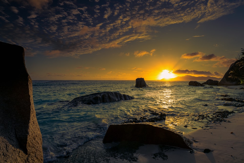 the sun is setting over the ocean with rocks in the foreground