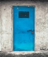 A bright blue metal door with a vent grid near the top, securely locked with a padlock. The door is set in a rough, textured gray concrete wall, suggesting an industrial or utilitarian setting.