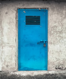 A bright blue metal door with a vent grid near the top, securely locked with a padlock. The door is set in a rough, textured gray concrete wall, suggesting an industrial or utilitarian setting.