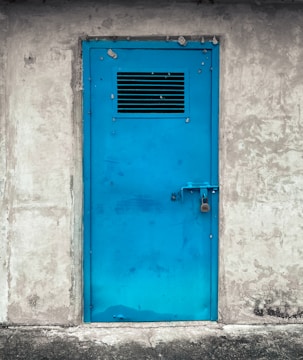 A bright blue metal door with a vent grid near the top, securely locked with a padlock. The door is set in a rough, textured gray concrete wall, suggesting an industrial or utilitarian setting.