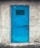 A bright blue metal door with a vent grid near the top, securely locked with a padlock. The door is set in a rough, textured gray concrete wall, suggesting an industrial or utilitarian setting.