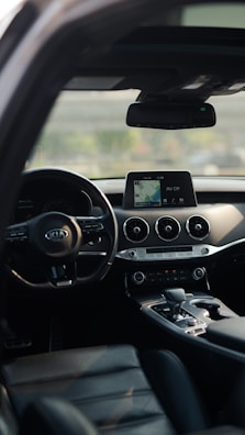 Interior of a car featuring a black dashboard and modern control panel with a digital screen displaying navigation and settings. The view includes air vents, a gear shift, and the steering wheel with a Kia logo. Sunlight subtly illuminates parts of the leather seats and interior.