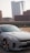A sleek gray Kia car is parked in a lot with an urban backdrop featuring modern high-rise buildings. The car's design is sporty, with distinctive alloy wheels and an aerodynamic shape. The setting appears to be a city environment with clear skies.