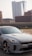 A sleek gray Kia car is parked in a lot with an urban backdrop featuring modern high-rise buildings. The car's design is sporty, with distinctive alloy wheels and an aerodynamic shape. The setting appears to be a city environment with clear skies.