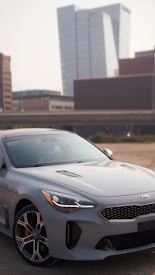 A sleek gray Kia car is parked in a lot with an urban backdrop featuring modern high-rise buildings. The car's design is sporty, with distinctive alloy wheels and an aerodynamic shape. The setting appears to be a city environment with clear skies.