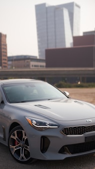 A sleek gray Kia car is parked in a lot with an urban backdrop featuring modern high-rise buildings. The car's design is sporty, with distinctive alloy wheels and an aerodynamic shape. The setting appears to be a city environment with clear skies.