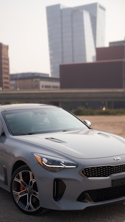 A sleek gray Kia car is parked in a lot with an urban backdrop featuring modern high-rise buildings. The car's design is sporty, with distinctive alloy wheels and an aerodynamic shape. The setting appears to be a city environment with clear skies.
