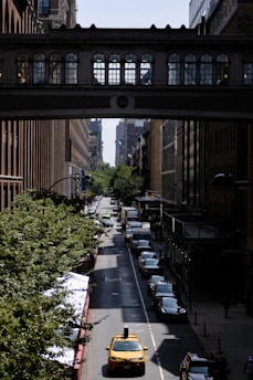A yellow taxi drives down a tree-lined city street flanked by tall buildings. An elevated bridge with ornate windows crosses above the street, creating a shadow. Several cars are lined up along the curb, and a view of distant city buildings is visible between the architectural structures.