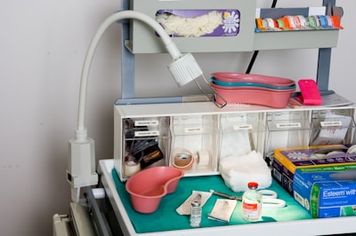 A medical supply cart featuring an adjustable lamp, various medical containers, and supplies organized neatly. Items include pink kidney trays, a container with gloves, rolls of tape, gauze pads, a pair of scissors, and bottles of medical liquids. The cart also holds boxes labeled for gloves and other medical essentials.