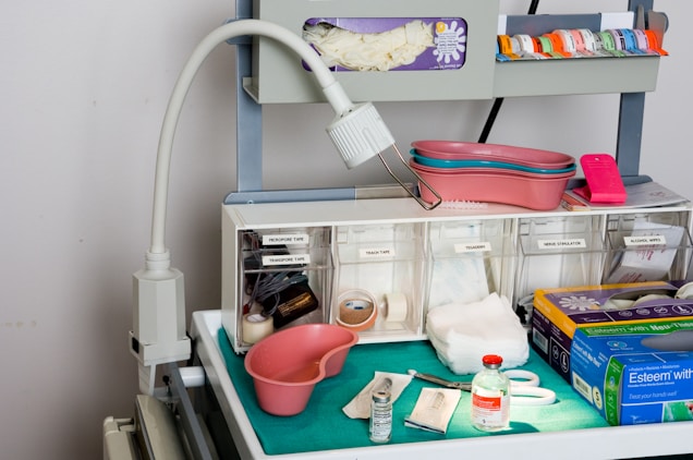 A medical supply cart featuring an adjustable lamp, various medical containers, and supplies organized neatly. Items include pink kidney trays, a container with gloves, rolls of tape, gauze pads, a pair of scissors, and bottles of medical liquids. The cart also holds boxes labeled for gloves and other medical essentials.