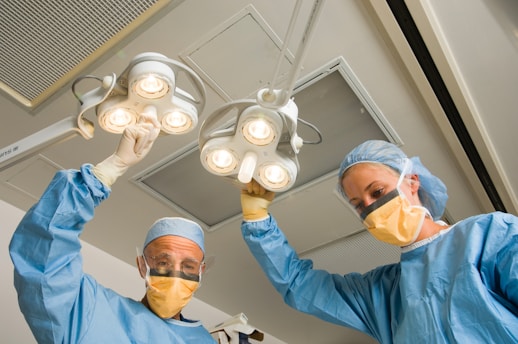 Two medical professionals wearing protective gear work in an operating room, adjusting overhead surgical lights. They are dressed in blue surgical gowns, caps, and yellow masks.