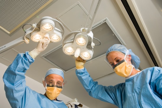 Two medical professionals wearing protective gear work in an operating room, adjusting overhead surgical lights. They are dressed in blue surgical gowns, caps, and yellow masks.