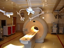 A hospital room containing an MRI machine surrounded by overhead lights and various medical equipment. The room has a clean and clinical environment with red cabinets on the left and diagnostic machines on the right.